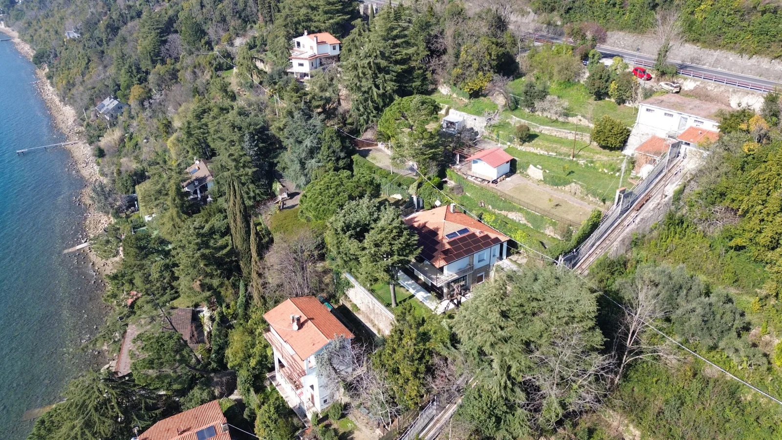 The plot seen from the drone: the terraces with the red-roofed shed, the neighbouring houses, the coast road and the sea on the left