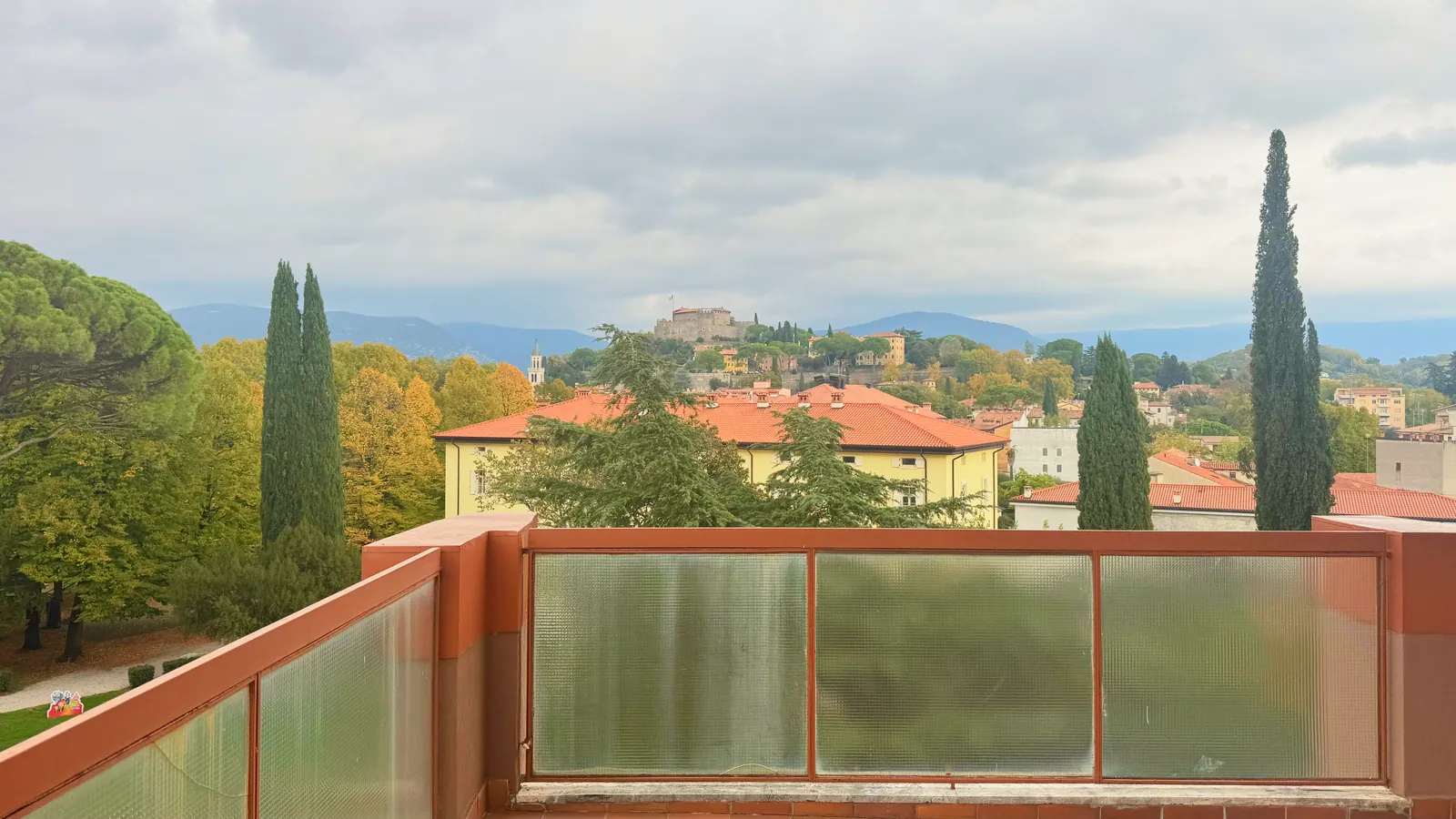 Dalla terrazza del quarto piano verso nord-est: in primo piano il parapetto, poi alberi e tetti, al centro il colle col Castello di Gorizia, dietro i monti sotto un cielo coperto
