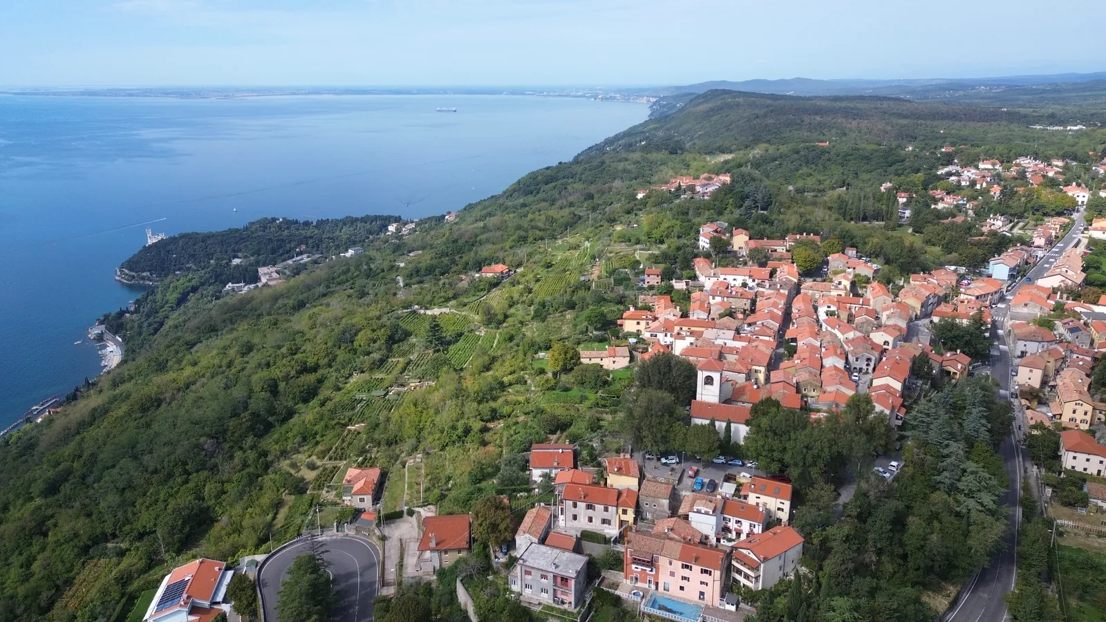 Contovello von oben: das Dorf am Hang, die Kirche San Gerolamo mit dem weißen Glockenturm, die Weinterrassen und links am Meer das Schloss Miramare.