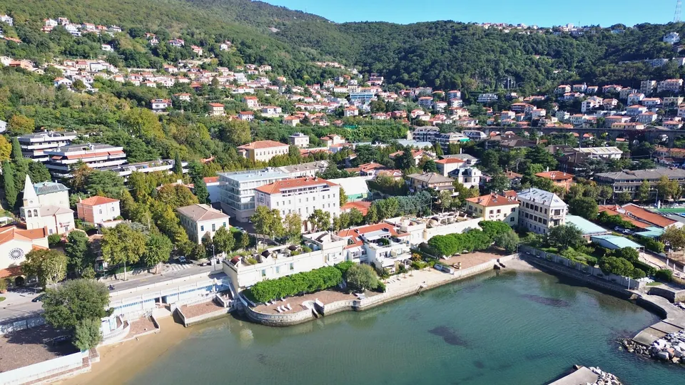 A small bay with a pale beach and green water, a promenade with low walls, villas and small apartment buildings among the trees on the slope.