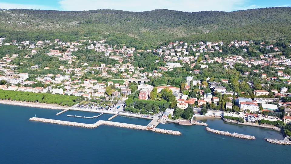 A long green shore seen from the sea: a row of pines on the left, a small harbour enclosed by a rock breakwater in the middle, pale houses climbing a wooded hill up to a ridge behind.