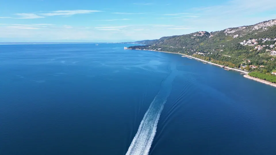 Calm blue sea crossed by the white wake of a motorboat; on the right the green coast and, in the distance, hills dropping to the water.