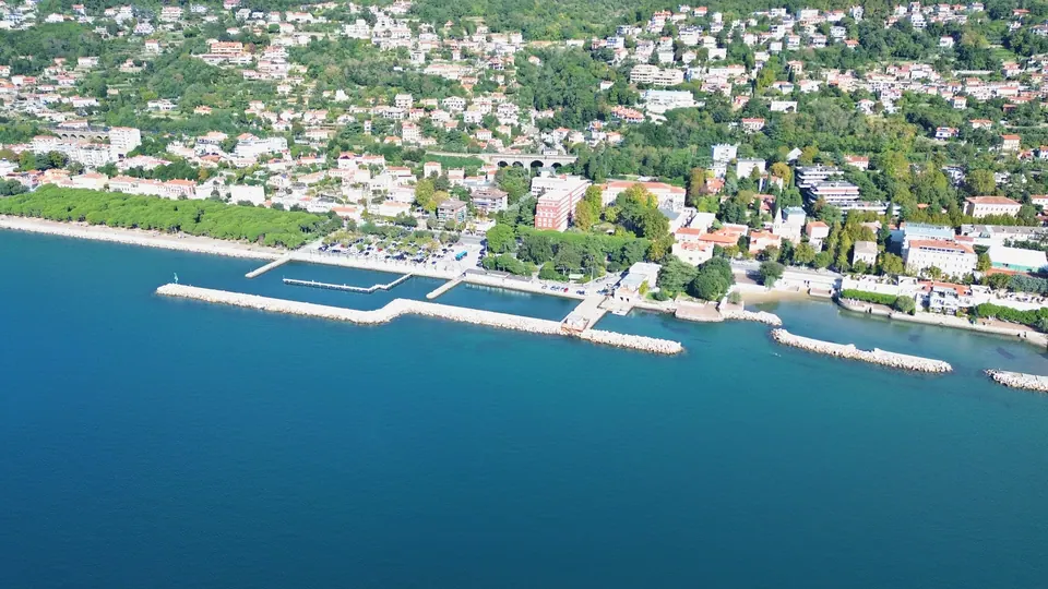 A small harbour enclosed by a long rock breakwater, with pontoons and boats; behind it a promenade, a row of pines on the left and houses on the hill.