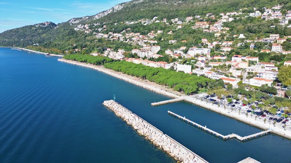 A small harbour with jetties and a rock breakwater; behind it, a seafront promenade with a long row of pines and, further on, the coast rising towards rocky slopes.