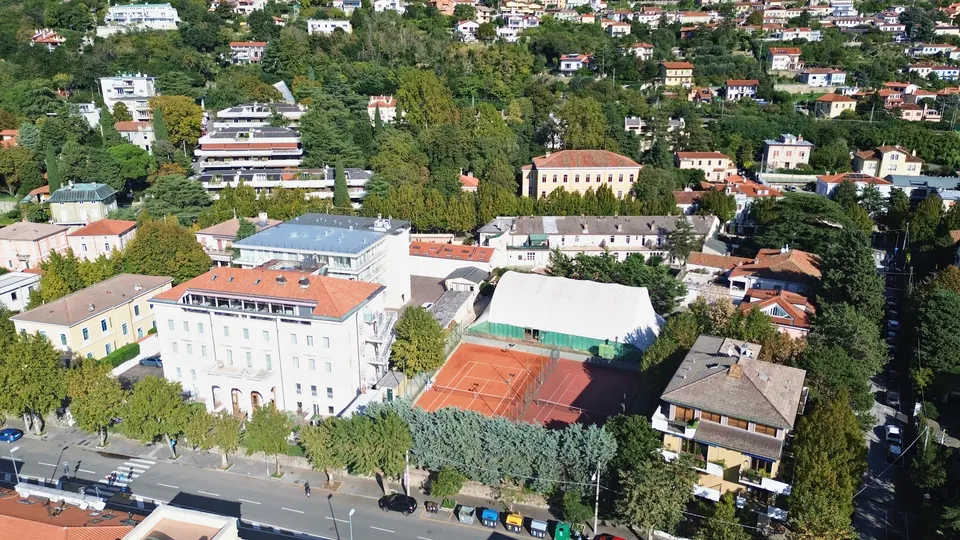 From above, red clay tennis courts next to a white building, among trees and small apartment blocks; below, a tree-lined road with parked cars.