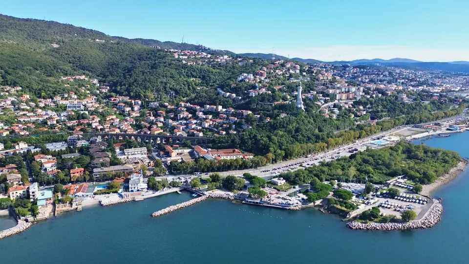 From above, a coastline leading to a city: on the right a flat point with trees and boats, on the hill a tall, slender white lighthouse, in the distance the city's buildings along the sea.