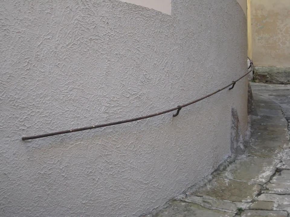 Un corrimano di ferro fissato al muro curvo di una casa, lungo una strada lastricata in salita