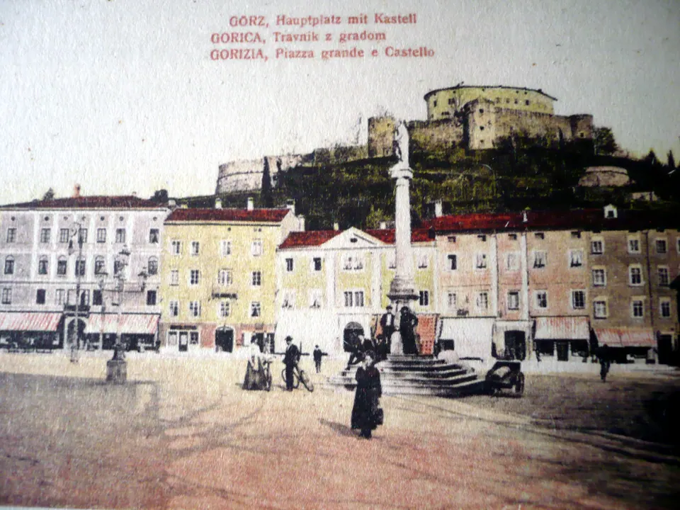 Cartolina colorata a mano: una piazza con una colonna sormontata da una statua, case a tre piani e, sul colle dietro, le mura del castello; in alto la scritta in tedesco, sloveno e italiano.