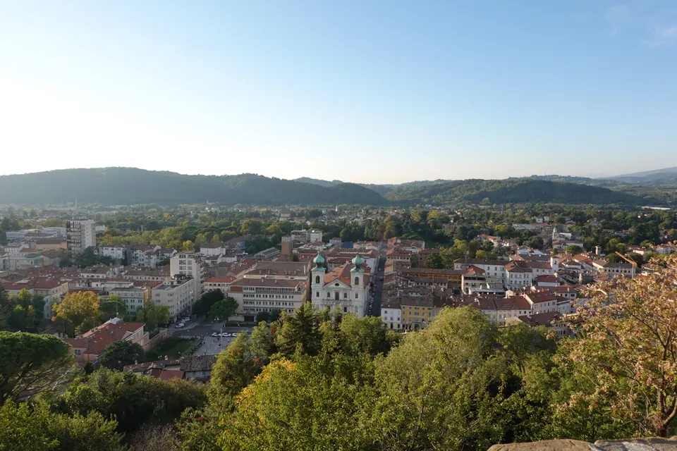 La città dall'alto: tetti rossi, al centro una chiesa con due torri dalle cupole verdi, e dietro colline boscose.