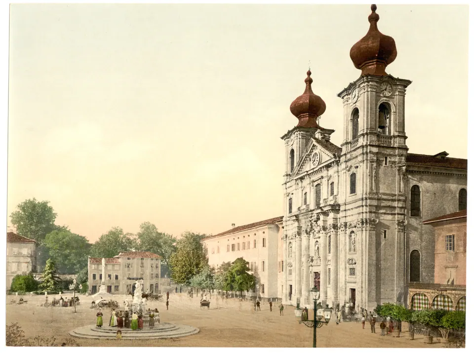 Veduta a colori di fine Ottocento: una grande piazza con una fontana al centro e, a destra, la facciata di una chiesa barocca con due torri a cipolla.