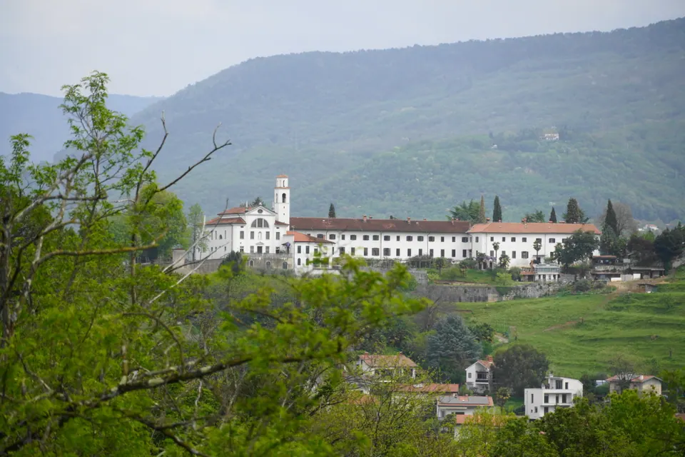 Un convento bianco con il campanile fra gli alberi di una collina verde, e dietro un monte boscoso.