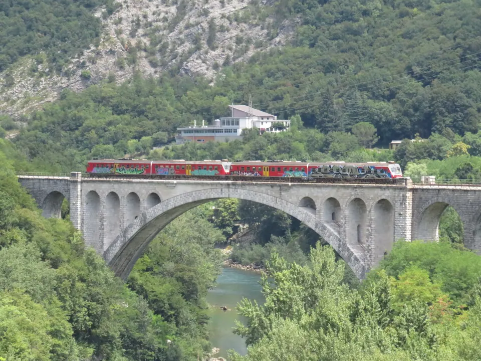 Un treno rosso passa su un ponte ferroviario di pietra con un grande arco sopra il fiume verde, fra pendii boscosi.
