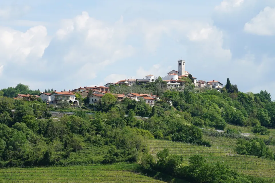 Un borgo con il campanile in cima a una collina, fra boschi e vigneti, sotto un cielo di nuvole bianche.