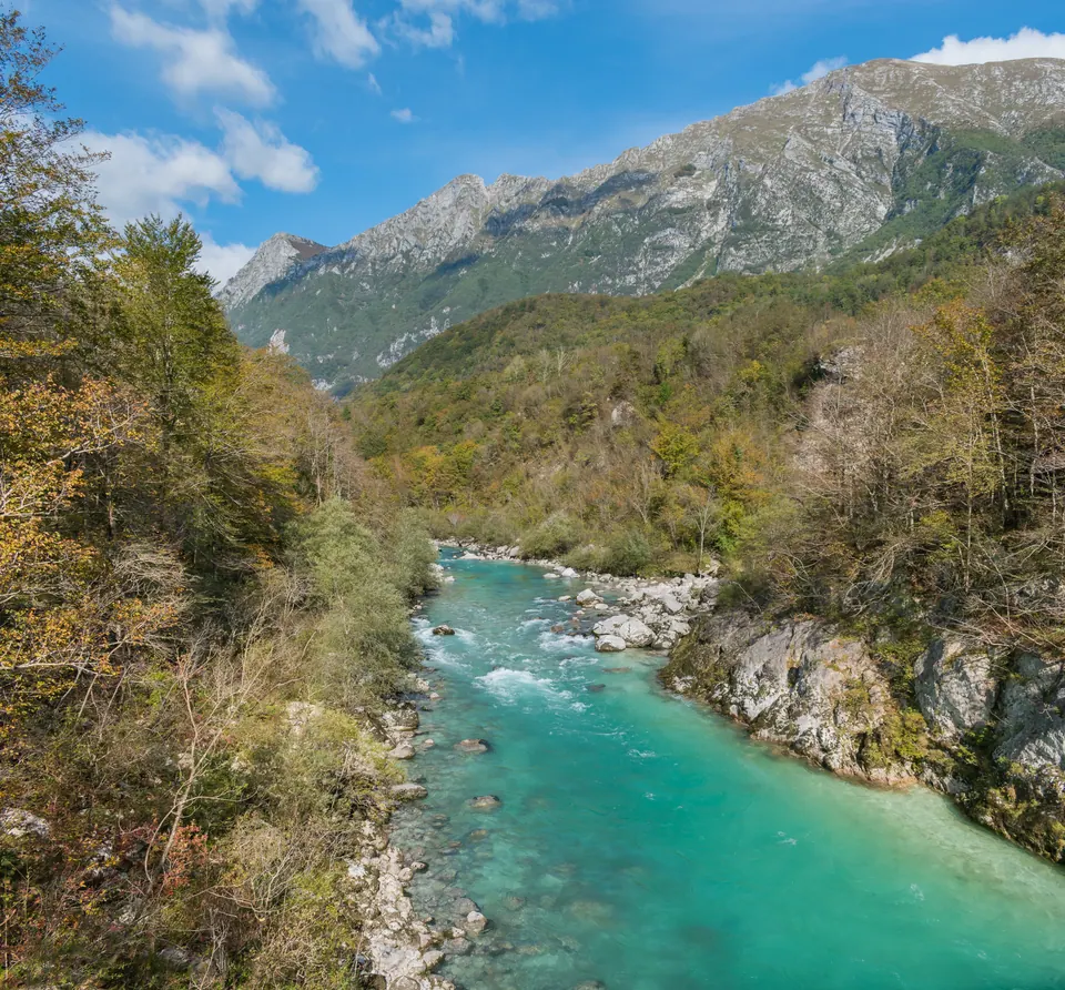 Un fiume color smeraldo scorre fra rocce chiare e boschi d'autunno, sotto montagne di roccia e un cielo azzurro.